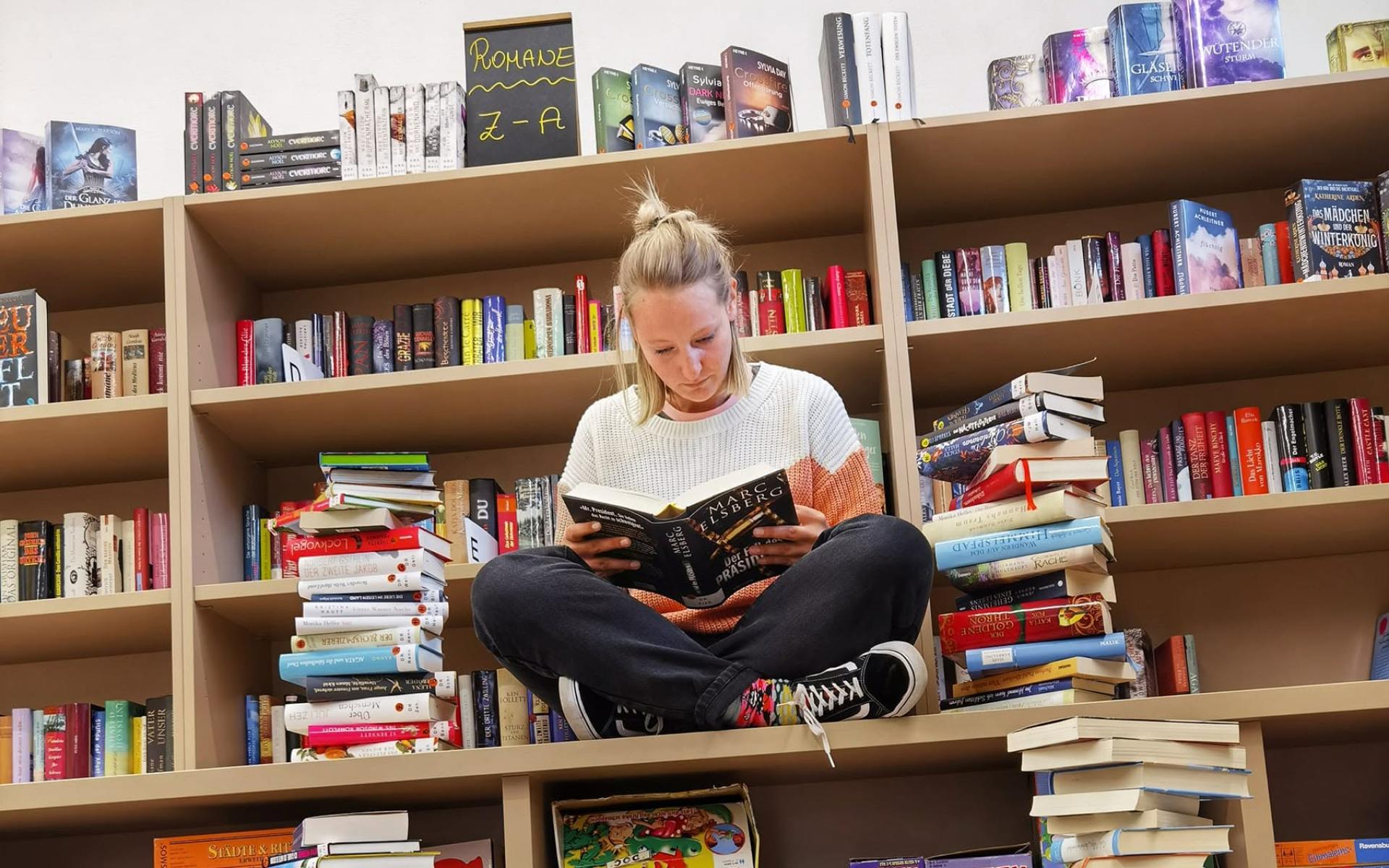 Frau sitzt im Schneidersitz zwischen Büchertürmen und Bücherregalen